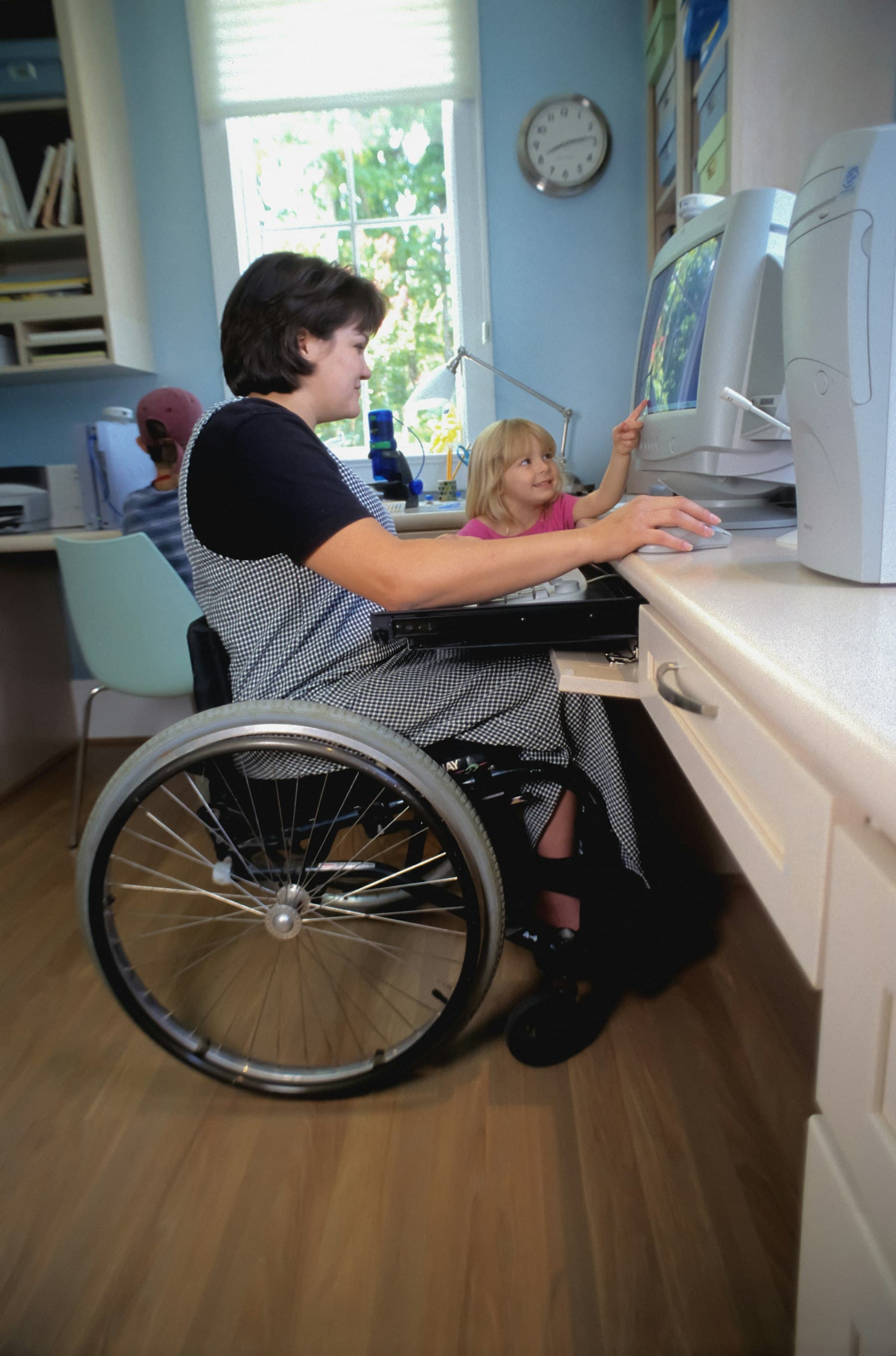 A woman who uses a wheelchair working at a computer at home while a young child looks on