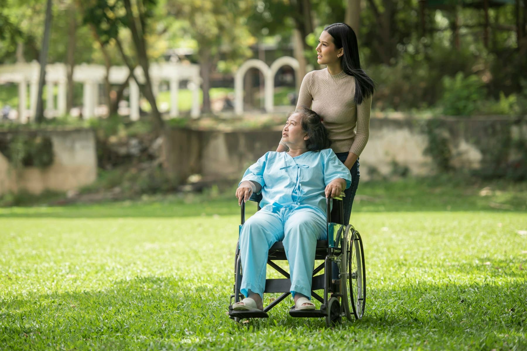 A woman walking alongside an older woman who uses a wheelchair in a sunny park